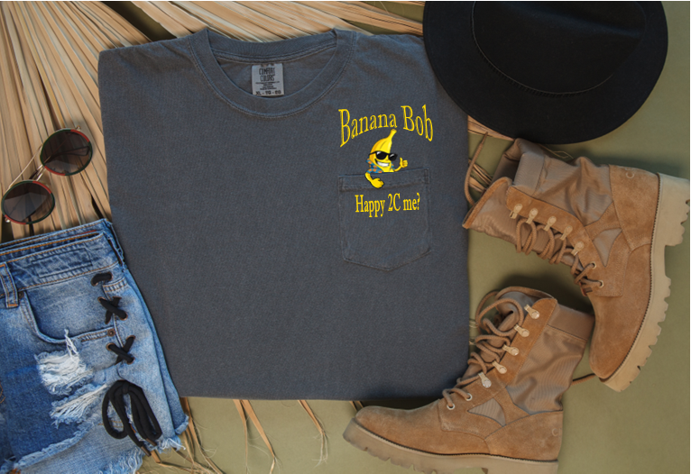 Gray pocket t-shirt with 'Banana Bob' Happy to see me? design, sunglasses, denim shorts, and boots on a wooden surface.