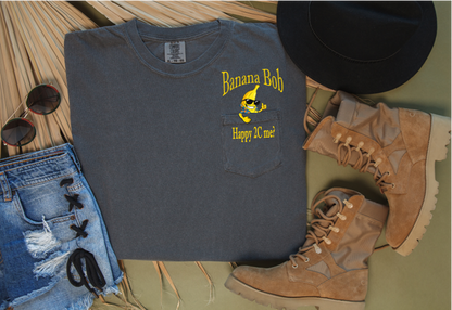 Gray pocket t-shirt with 'Banana Bob' Happy to see me? design, sunglasses, denim shorts, and boots on a wooden surface.